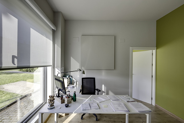 Bureau à domicile lumineux avec fenêtre, stores déroulants, murs gris/verts. Documents sur un grand bureau blanc, chaise, ordinateur.