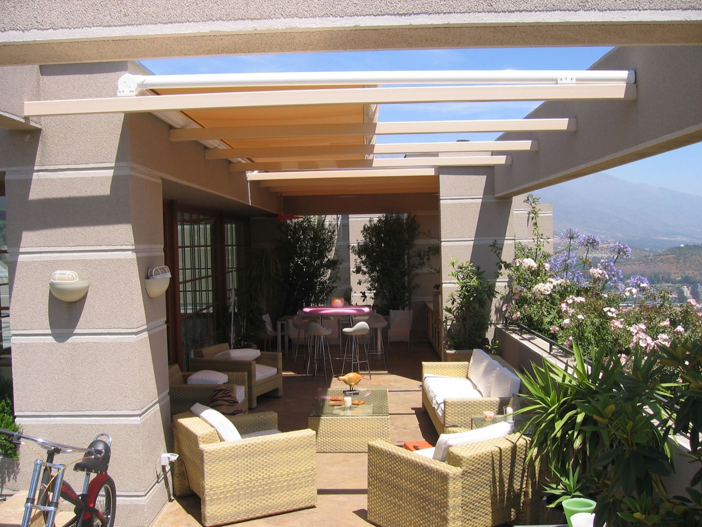 Terrasse moderne avec salon extérieur, store rétractable beige, plantes luxuriantes et vue sur la ville et la montagne.
