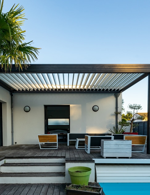 Terrasse bois avec pergola bioclimatique a lames orientables, mobilier de jardin et piscine, sous ciel bleu.