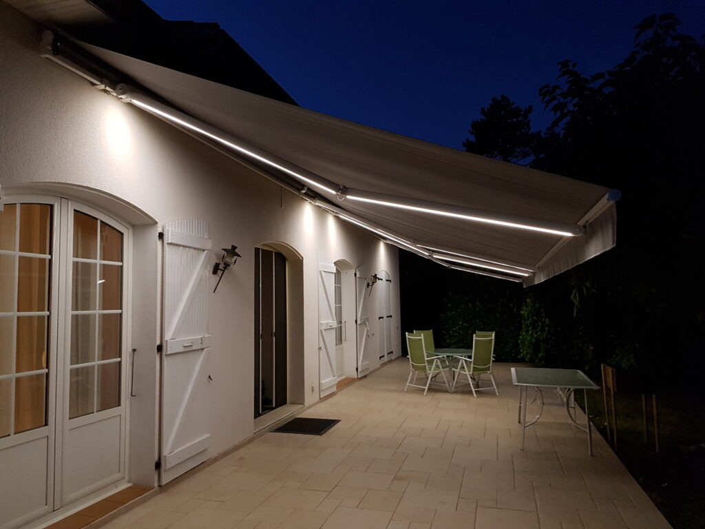 Façade de maison blanche la nuit, store banne avec éclairage LED intégré, terrasse carrelée, mobilier jardin.