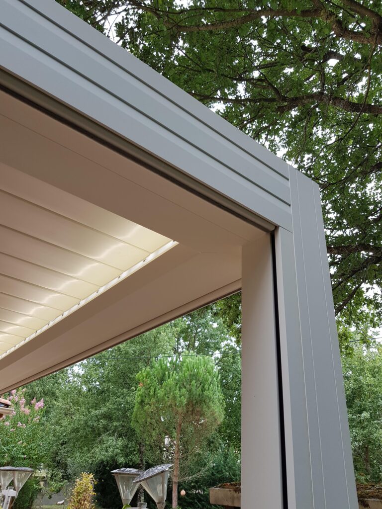 Pergola moderne en aluminium blanc avec toit rétractable beige. Structure sur terrasse avec vue sur jardin verdoyant et arbres.