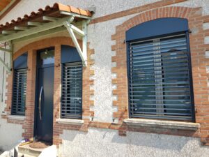 Façade maison avec porte sombre, 2 fenêtres grillagées, arches en briques, auvent bois et tuiles
