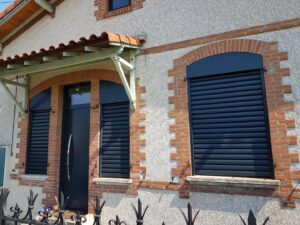 Façade maison avec porte noire, volets roulants noirs sur fenêtres à arches en brique. Auvent tuilé, mur texturé, clôture fer.