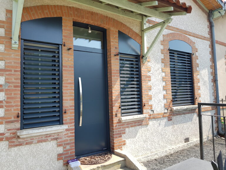 Façade maison: porte grise moderne, volets persiennes, arcades brique. Boîte Omfy sur marche d'escalier.