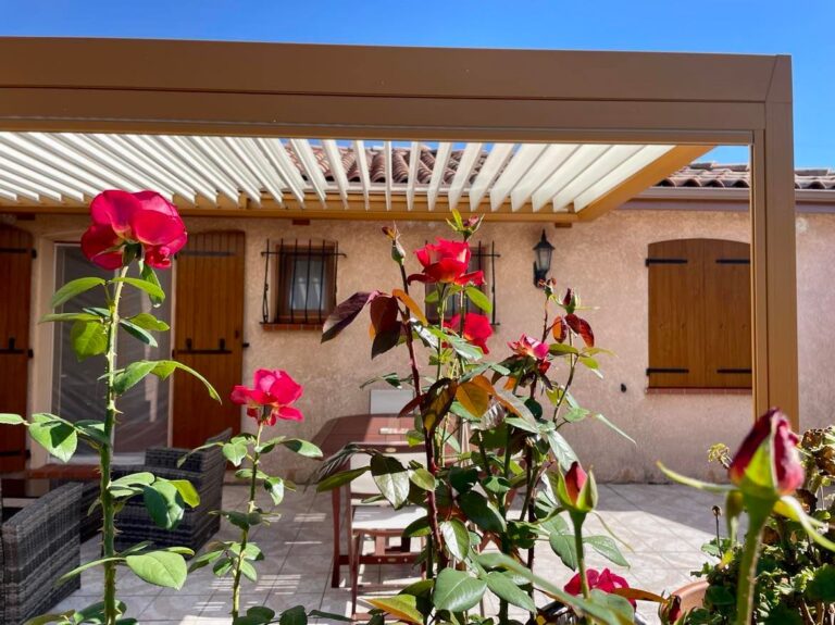 Patio ensoleillé avec pergola bioclimatique, rosiers rouges fleuris, maison aux volets bois et fenêtres grillagées.