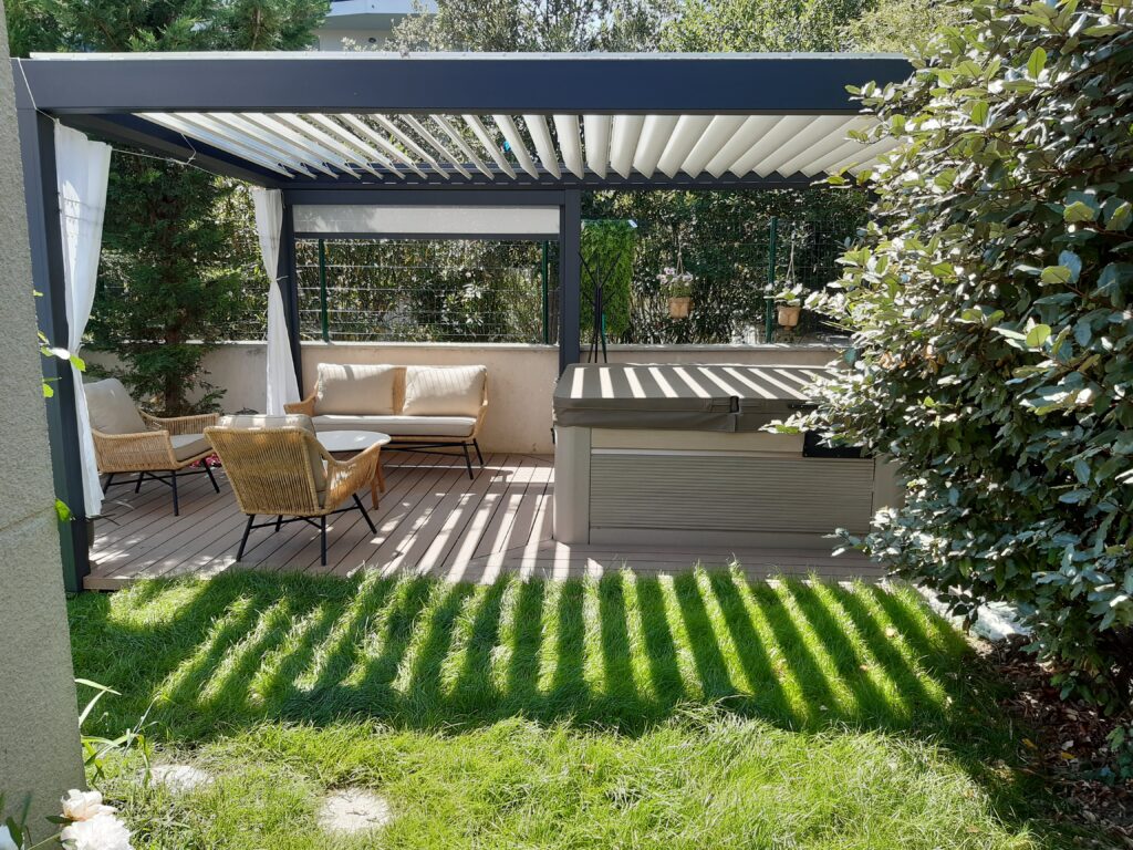 Terrasse bois avec pergola bioclimatique grise et rideaux blancs, abritant un salon de jardin en rotin et un spa couvert. Ombre sur l'herbe.