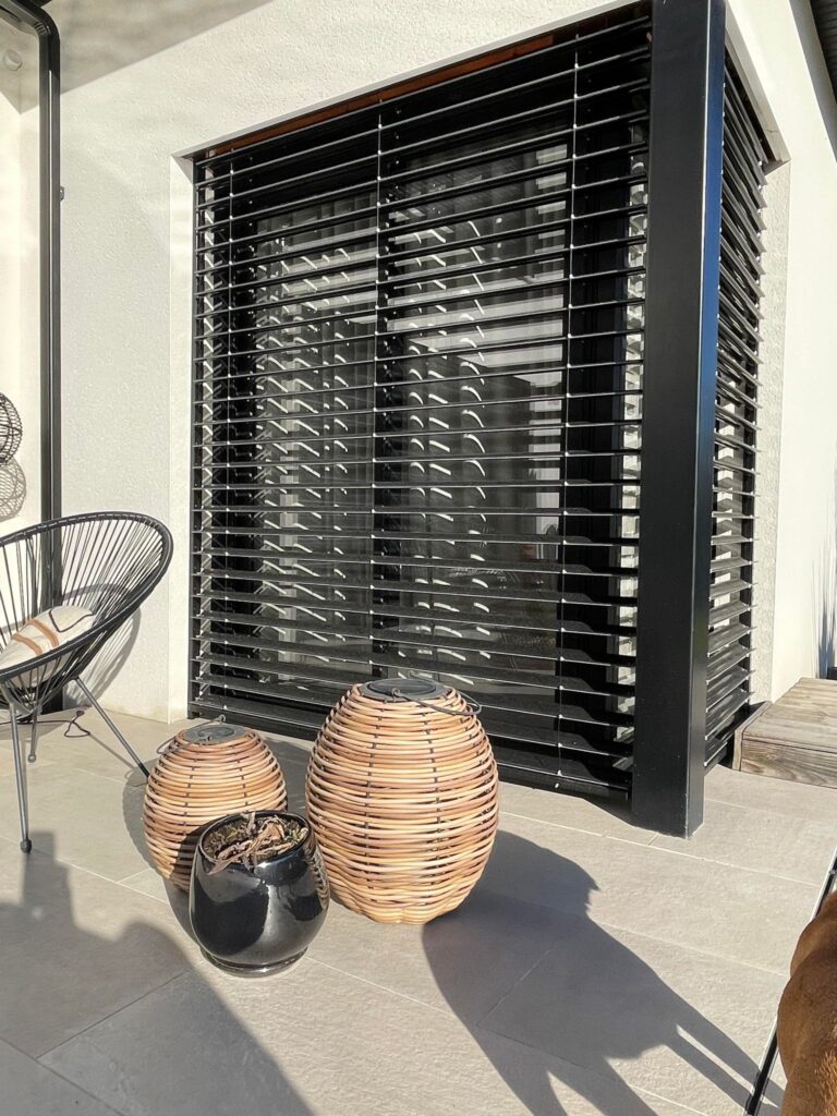 Façade blanche, large fenêtre avec brise-soleil noir, terrasse moderne carrelée, fauteuil, lampes décoratives tressées.