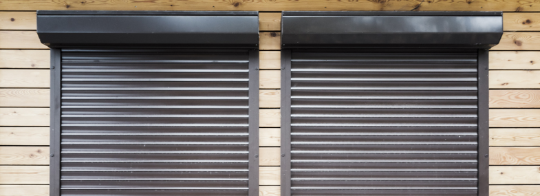 Deux volets roulants marron fermés sur une façade en bois clair. Encadrement et mécanismes visibles.