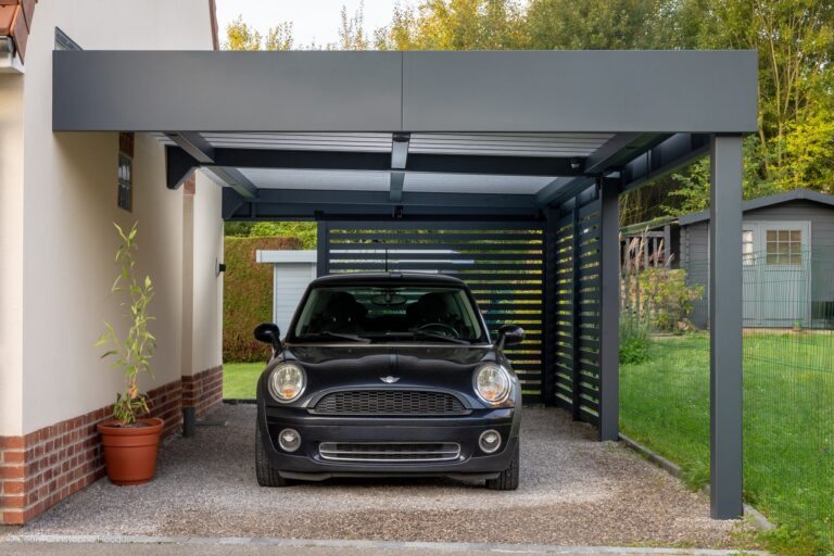 Mini Cooper noire sous un carport moderne gris adossé à une maison. Allée en gravier, écran ajouré, jardin.