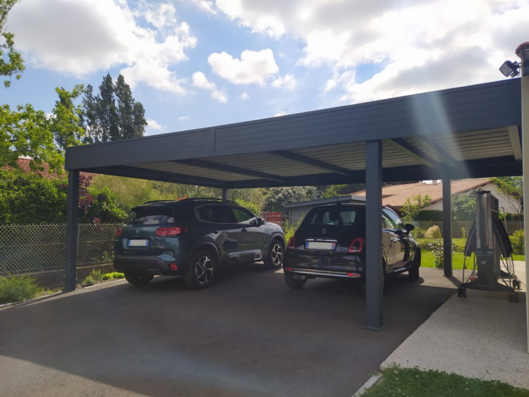 Carport moderne gris couvrant un SUV vert et une citadine noire. Jardin et clôture, ciel bleu nuageux.