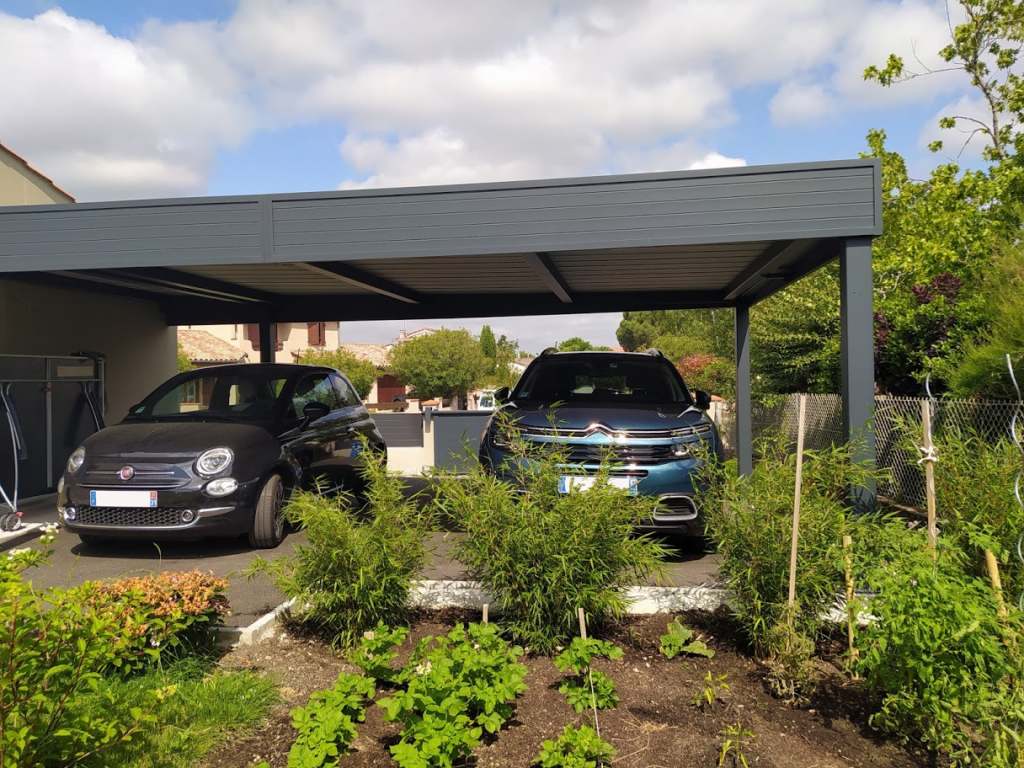 Deux voitures (Fiat 500, Citroën) garées sous un abri gris, avec un jardin au premier plan sous un ciel nuageux.