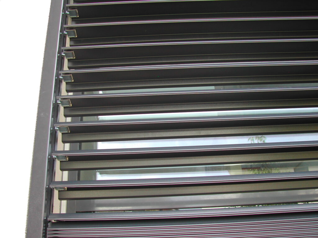 Store Extérieur à Lames Orientables - Brise-Soleil Design Store extérieur sombre à lames horizontales orientables, montrant le mécanisme latéral et des reflets de l'environnement.