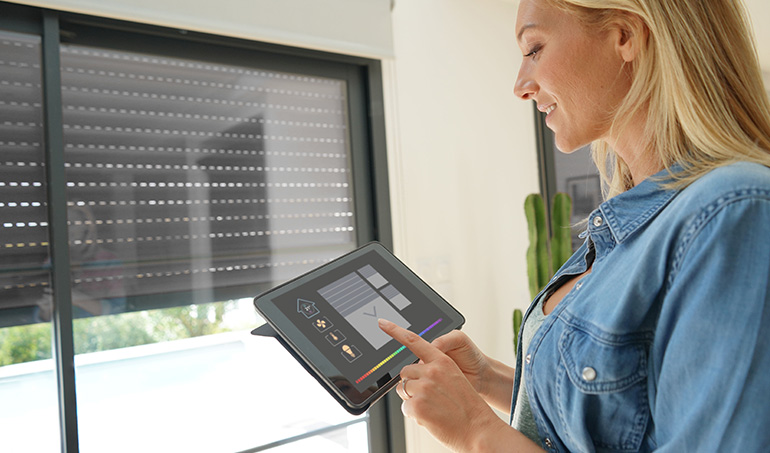 Femme souriante utilise une tablette pour contrôler des volets roulants intelligents sur une grande fenêtre.