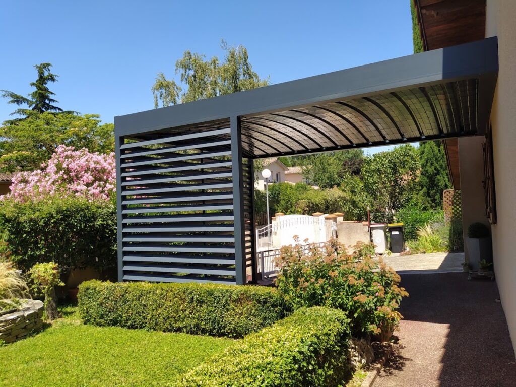 Pergola moderne grise adossée à une maison, avec toit transparent voûté et paroi à lattes horizontales, dans un jardin verdoyant et fleuri.