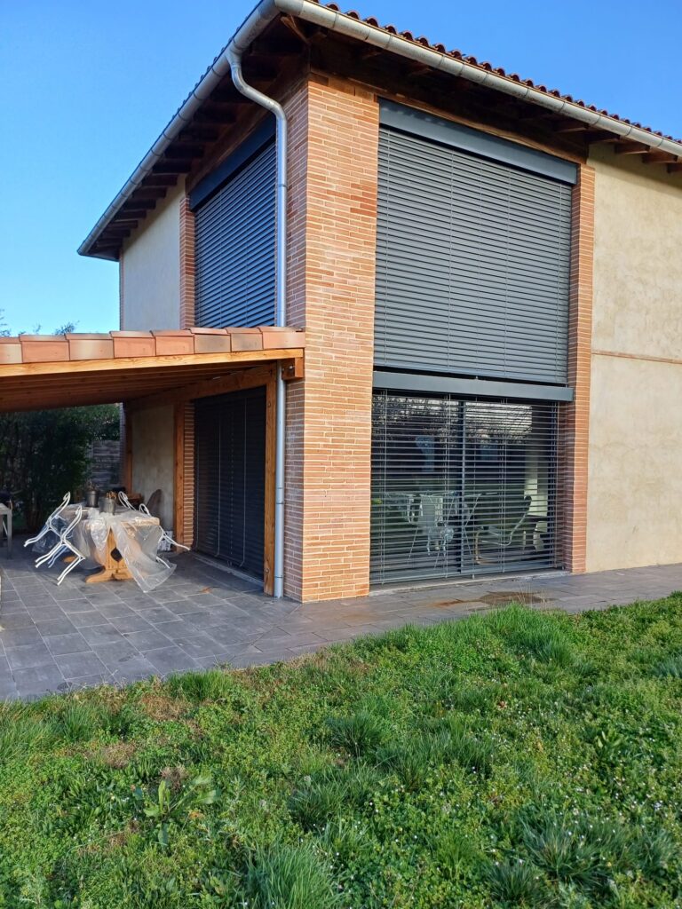 Maison moderne avec volets roulants foncés sur grandes fenêtres, terrasse couverte avec table et chaises, carrelage gris, jardin.