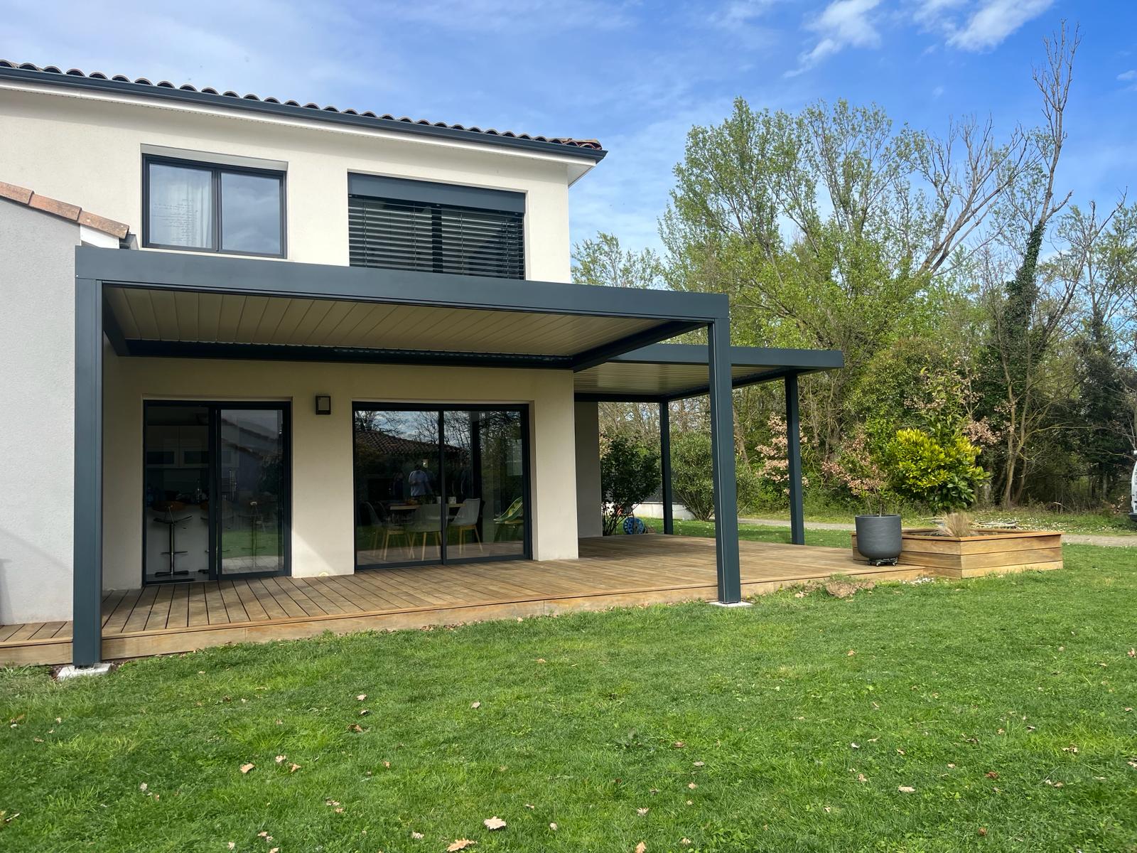Maison moderne avec pergola anthracite couvrant une terrasse en bois. Jardin vert avec arbres et ciel bleu clair.
