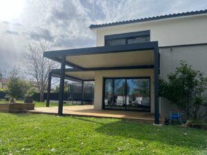 Maison moderne beige et grise avec pergola noire, grande terrasse bois et jardin arboré avec trampoline.