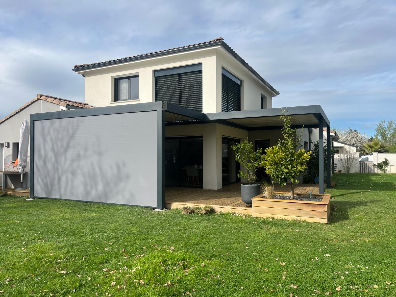 Maison moderne avec terrasse bois, pergola bioclimatique grise à toit rétractable et écran vertical, dans jardin pelouse.