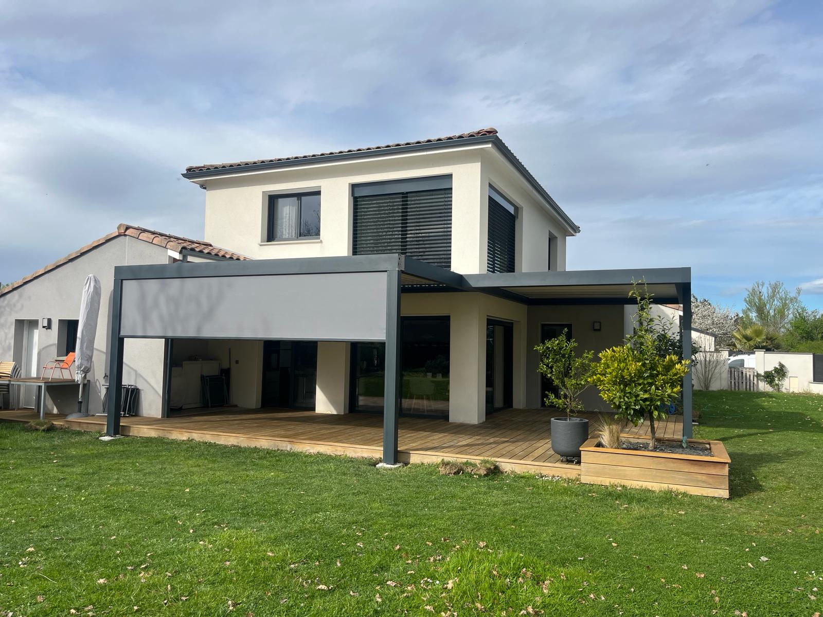 Maison moderne avec terrasse en bois et pergola grise. La pergola est équipée d'un store rétractable gris. Jardin verdoyant avec plantes.