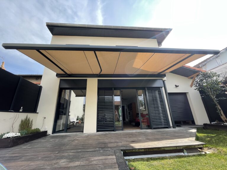Façade de maison moderne avec terrasse bois, grand auvant rétractable beige. Portes vitrées coulissantes, volets noirs et roulants.