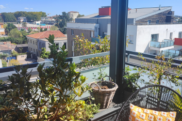 Vue depuis un balcon avec plantes, chaise et coussin, surplombant un paysage urbain de bâtiments et arbres sous un ciel bleu clair.