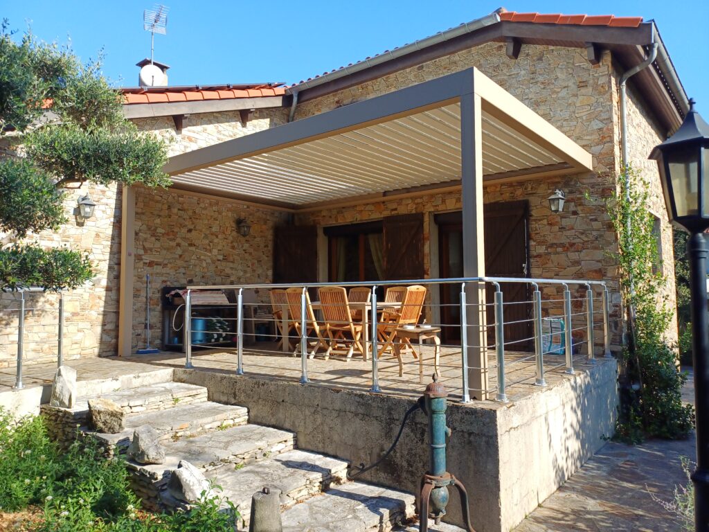 Maison en pierre traditionnelle avec terrasse couverte par pergola, meublée, escalier en pierre, balustrade, pompe vintage, jardin ensoleillé.