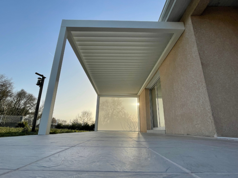 Pergola blanche moderne couvrant une terrasse avec dalles grises, vue sur le soleil couchant et arbres derrière un écran.