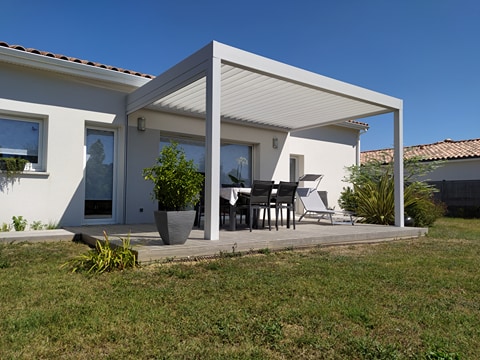 Pergola bioclimatique blanche sur terrasse bois devant une maison, avec table, chaises et transat dans le jardin ensoleillé.