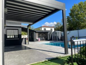 Espace extérieur moderne avec piscine bleue, pergola grise, pool house et patio carrelé sous ciel bleu.