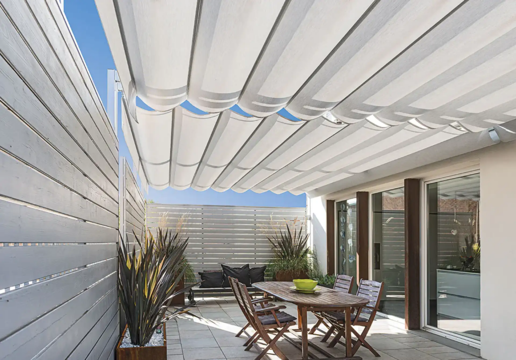 Terrasse ensoleillée avec store retractable blanc offrant de l'ombre sur une table de jardin en bois, des plantes et une clôture.
