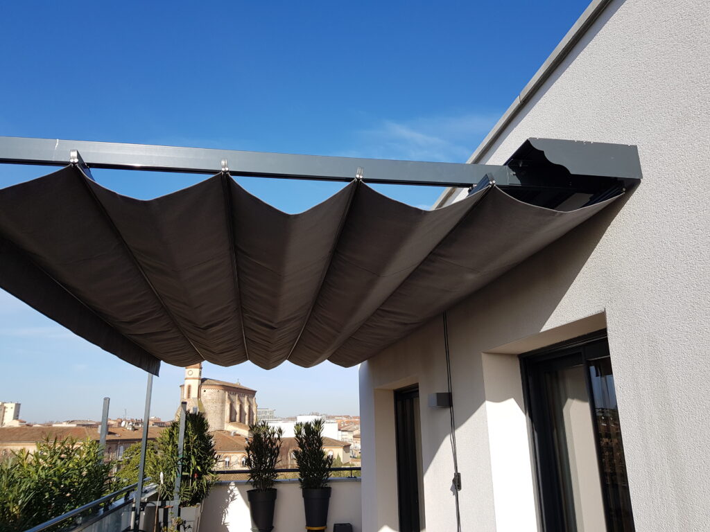 Store rétractable gris sombre déplié sur une terrasse, vue sur les toits de la ville, une église et ciel bleu.