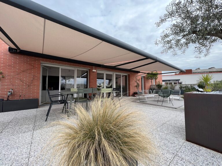 Terrasse moderne avec store banne beige, mobilier de jardin gris et vert, mur en briques rouges et plantes. Espace exterieur amenage.