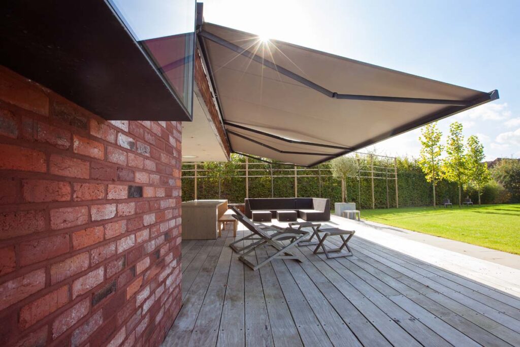 Grand store banne beige au-dessus d'une terrasse en deck bois avec mobilier extérieur près d'un mur en briques sous un soleil éclatant.