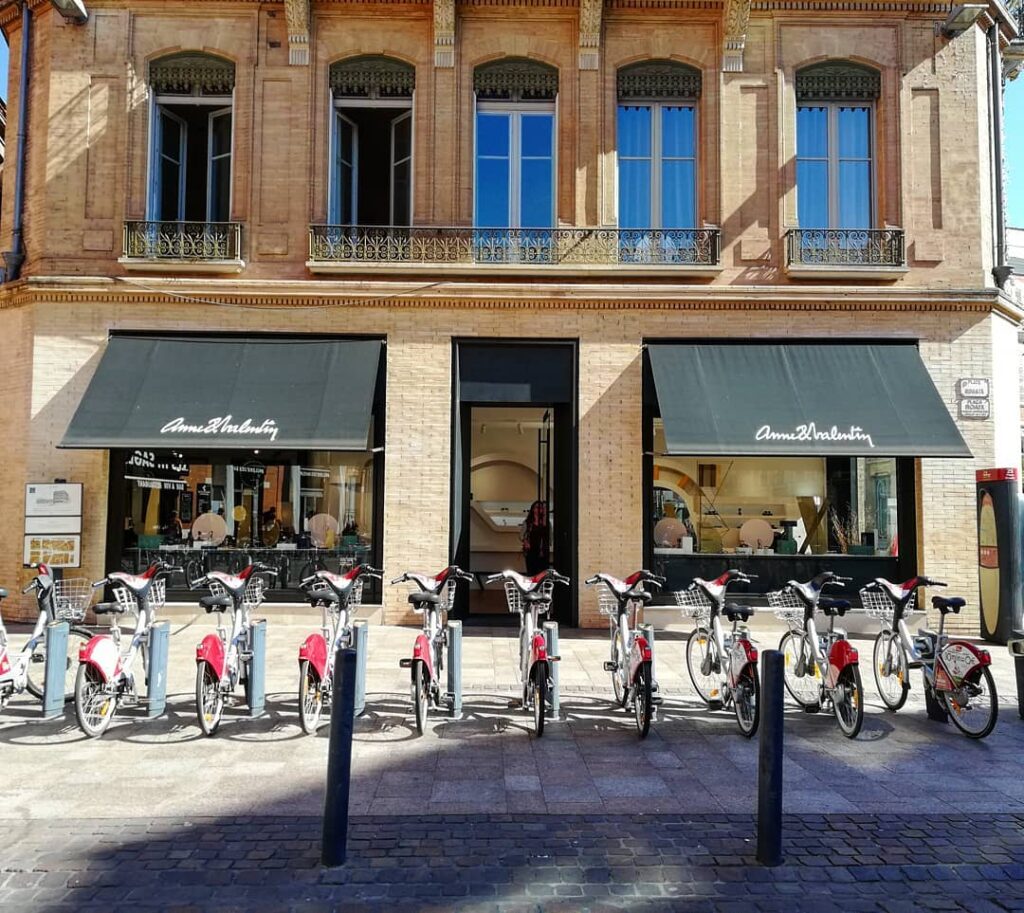 Façade du magasin Anne&Valentin avec marquise noire. De nombreux vélos rouges en libre-service sont garés devant sur le trottoir. Bâtiment en briques.