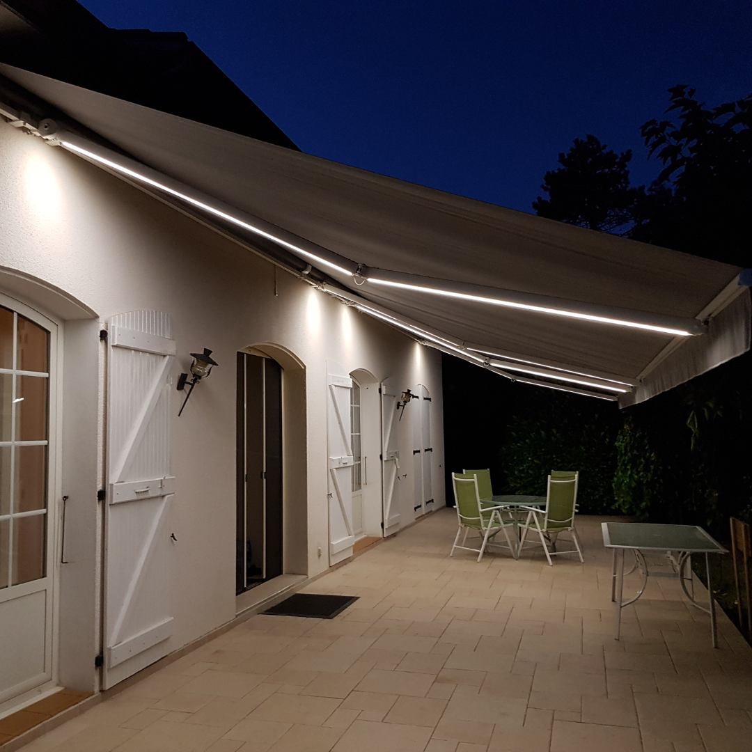Patio de maison éclairé la nuit par un store banne avec bande LED intégrée. Terrasse carrelée avec salon de jardin et table. Façade blanche avec volets.