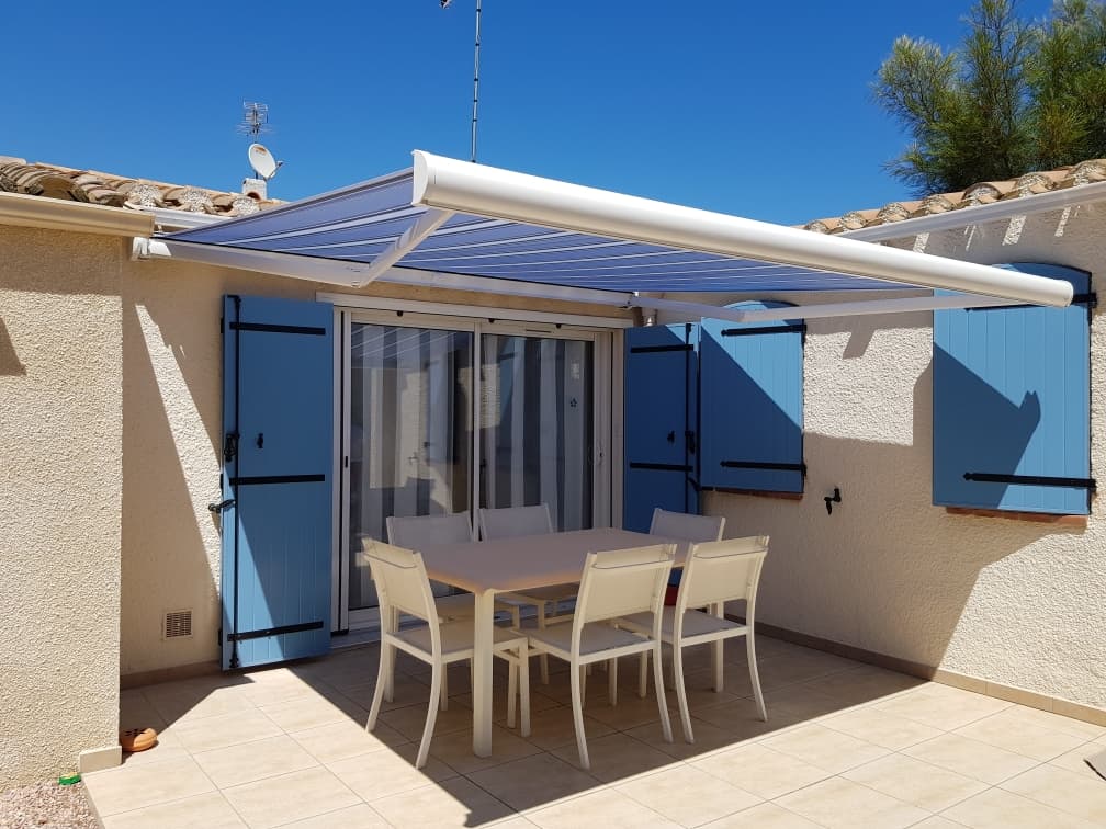 Terrasse ensoleillée avec un grand auvent bleu translucide couvrant une table et des chaises blanches. Façade beige avec volets bleus.