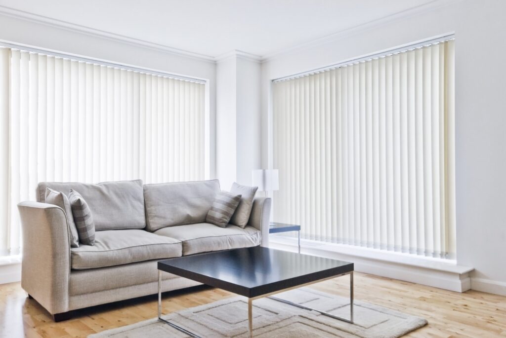 Salon moderne lumineux avec canapé gris, table basse noire, stores verticaux blancs. Parquet clair et tapis texturé.