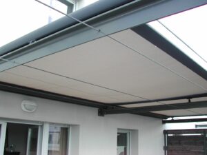 Store banne rétractable beige sur structure métallique foncée et câbles de guidage, installé au-dessus de fenêtres sur un mur blanc.