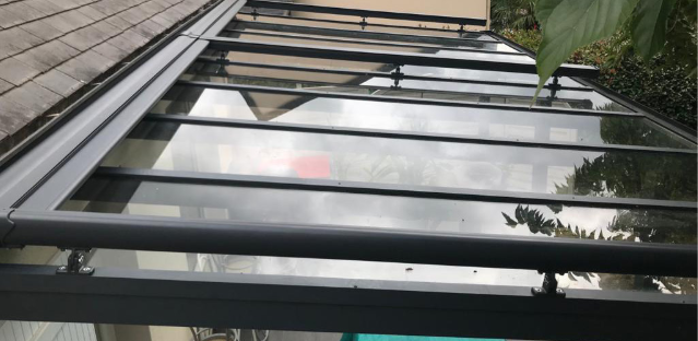 Verrière ou auvent en verre et métal gris foncé fixé à un toit de tuiles, avec des reflets du ciel et des arbres.