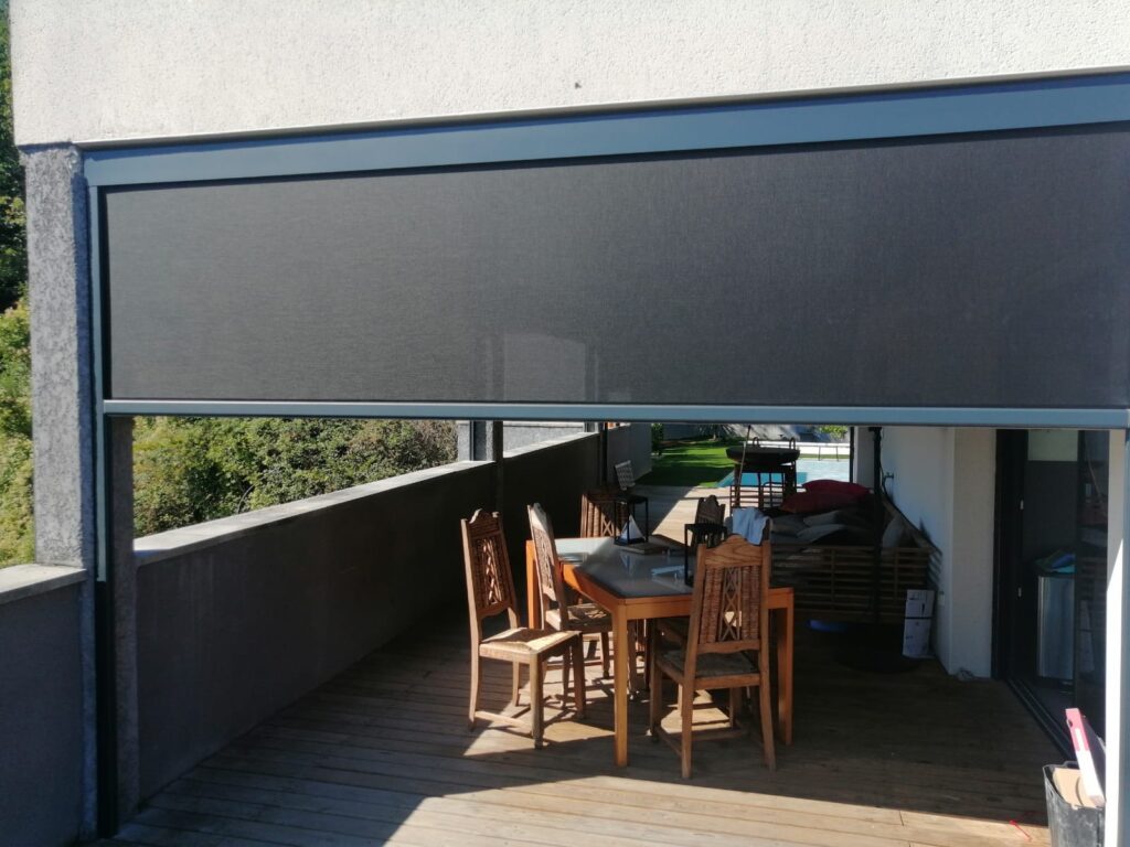 Store extérieur gris étendu sur une terrasse en bois avec table, chaises et vue piscine.