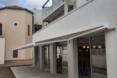 Bâtiment moderne avec grande baie vitrée, long store banne blanc sur terrasse. À côté, ancien bâtiment beige. Travaux d'aménagement extérieur.