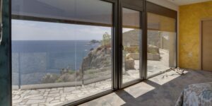 Baie vitrée vue mer et côte rocheuse depuis une chambre. Terrasse en pierre, soleil et ombres.