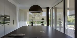 Cuisine moderne avec îlot central sombre, luminaire suspendu design, et large fenêtre avec stores semi-opaques. Vue sur jardin.