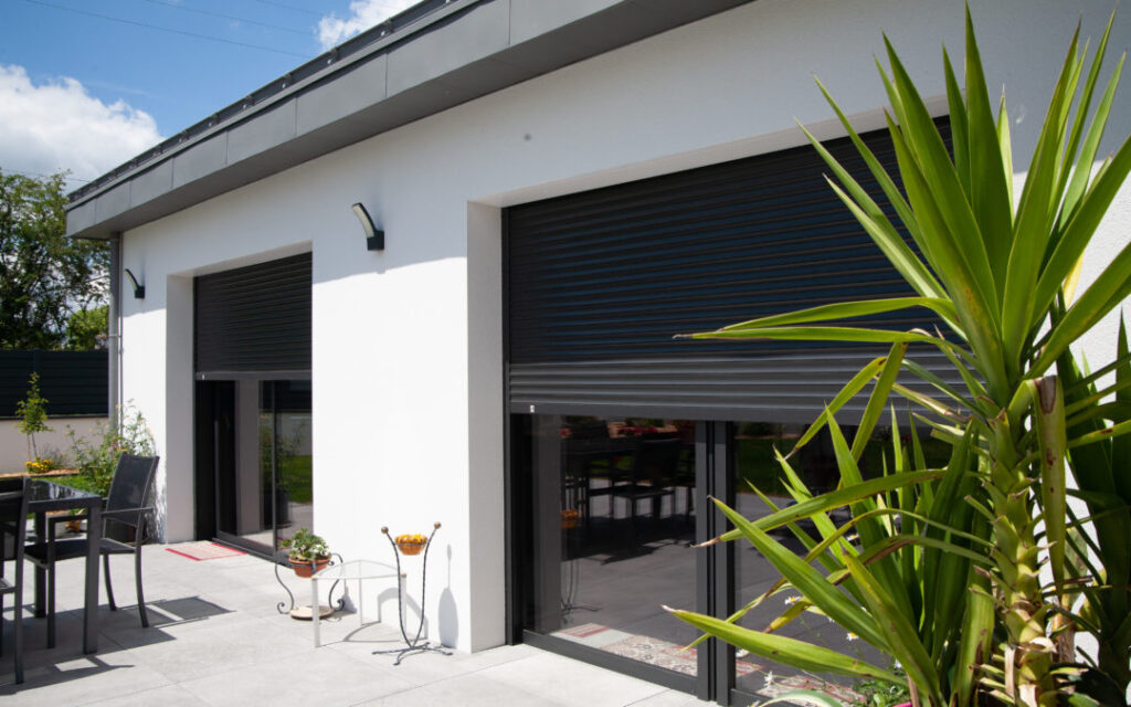 Maison moderne blanche avec deux grandes baies vitrées équipées de volets roulants noirs. Terrasse aménagée.