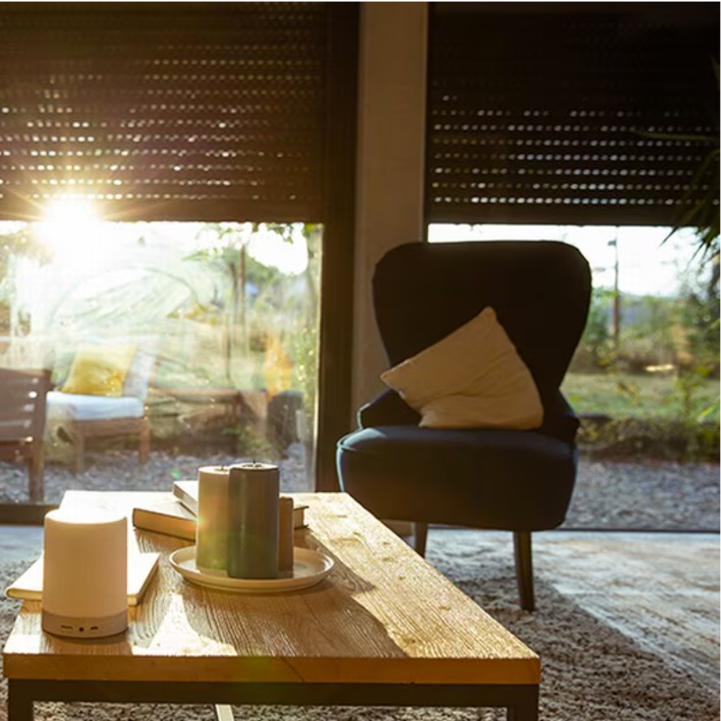 Salon lumineux avec volets roulants sombres baissés, soleil filtrant, fauteuil bleu, table en bois. Ambiance chaleureuse.