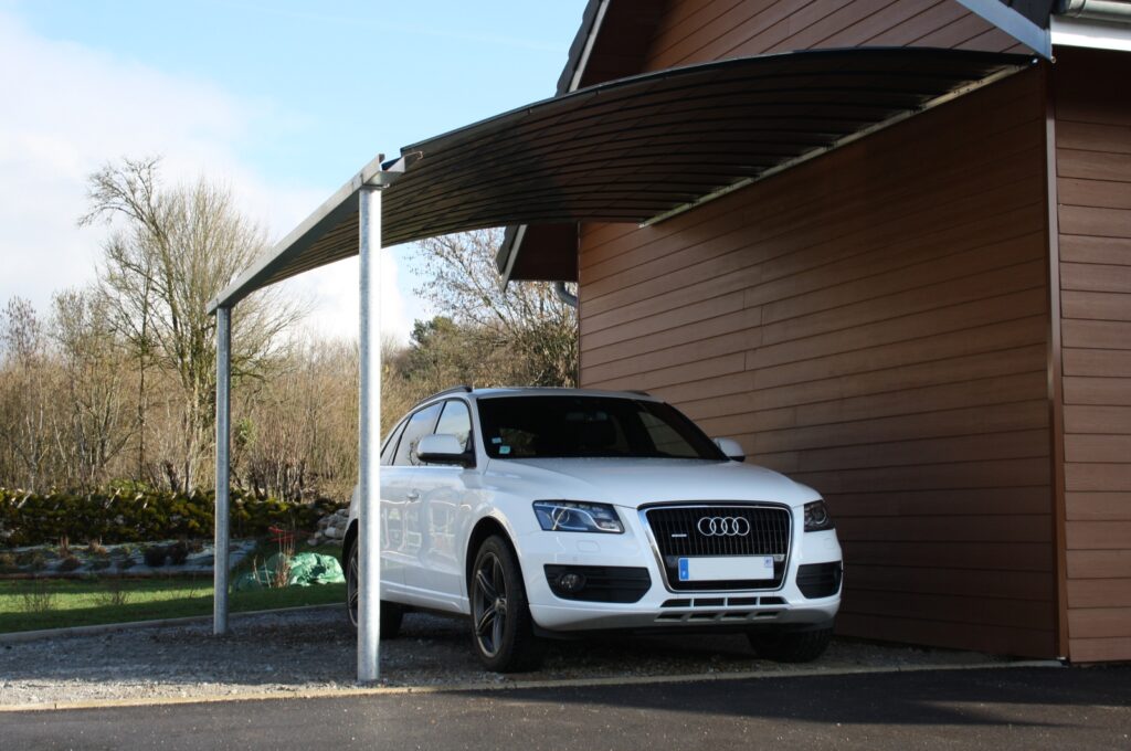 Audi Q5 blanc garé sous un carport moderne sombre adossé à une maison en bois marron.