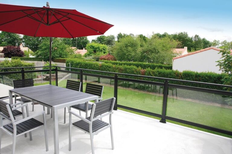 Balcon aménagé avec table grise, chaises, parasol rouge, garde-corps verre et aluminium, vue jardin.