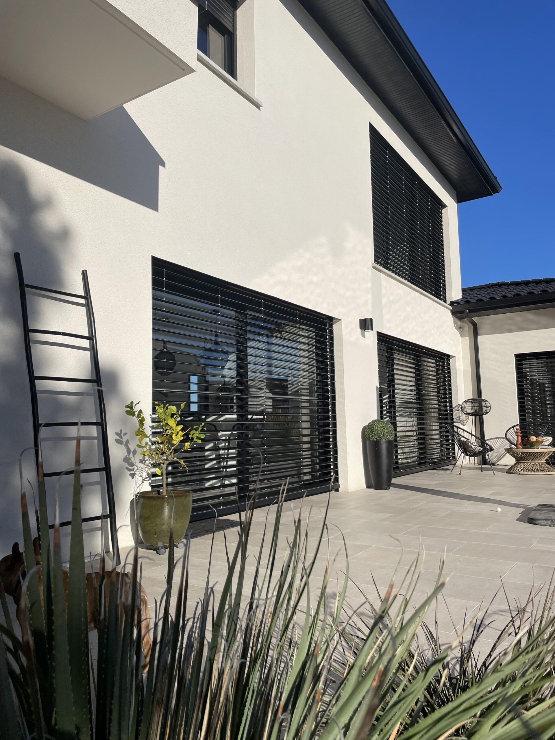 Maison blanche, stores noirs, grande baie vitrée, terrasse carrelée, plantes en pot, échelle noire, mobilier extérieur, ciel bleu ensoleillé.