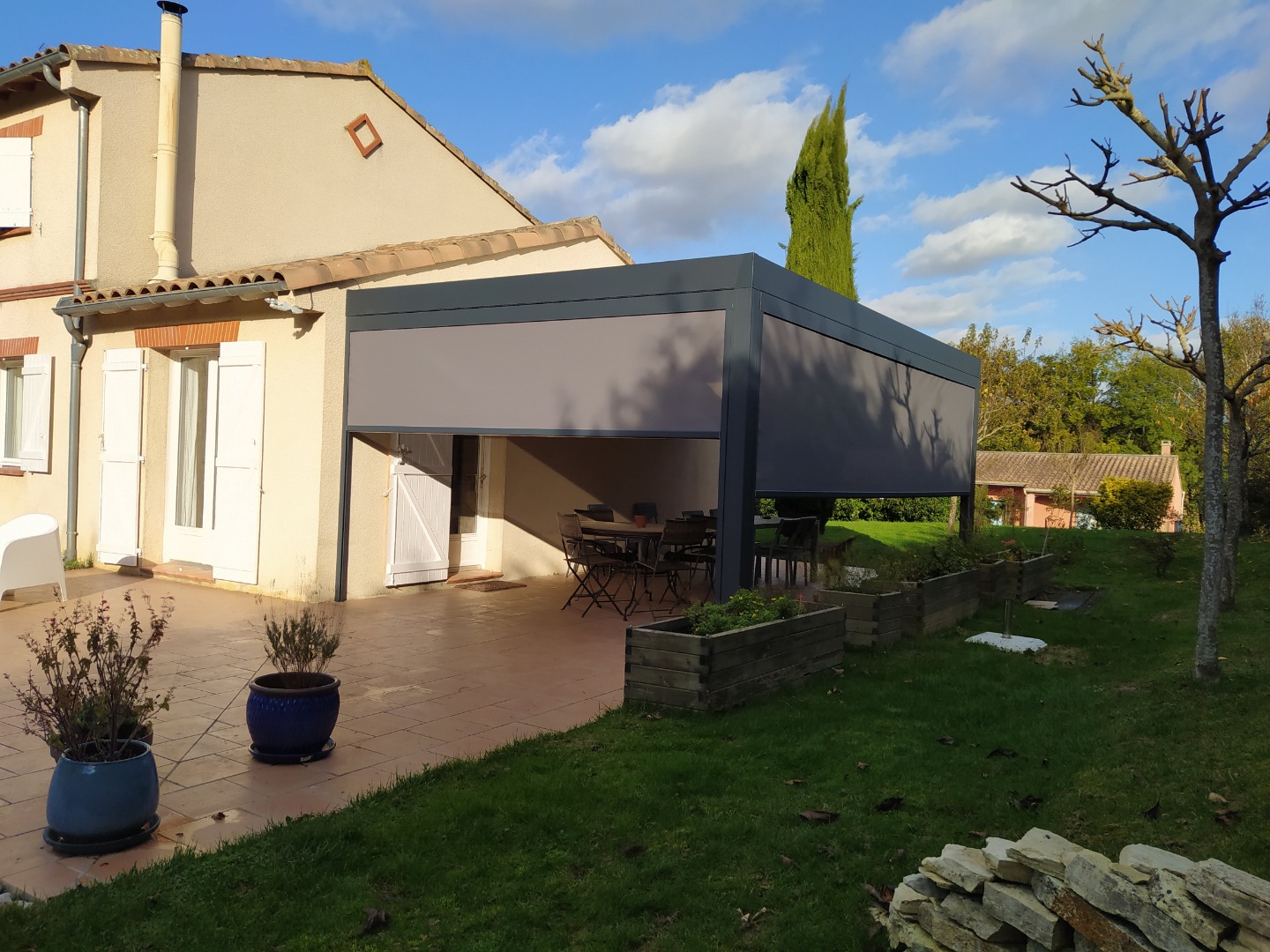Pergola bioclimatique grise avec stores attenante à une maison beige sur une terrasse ensoleillée.