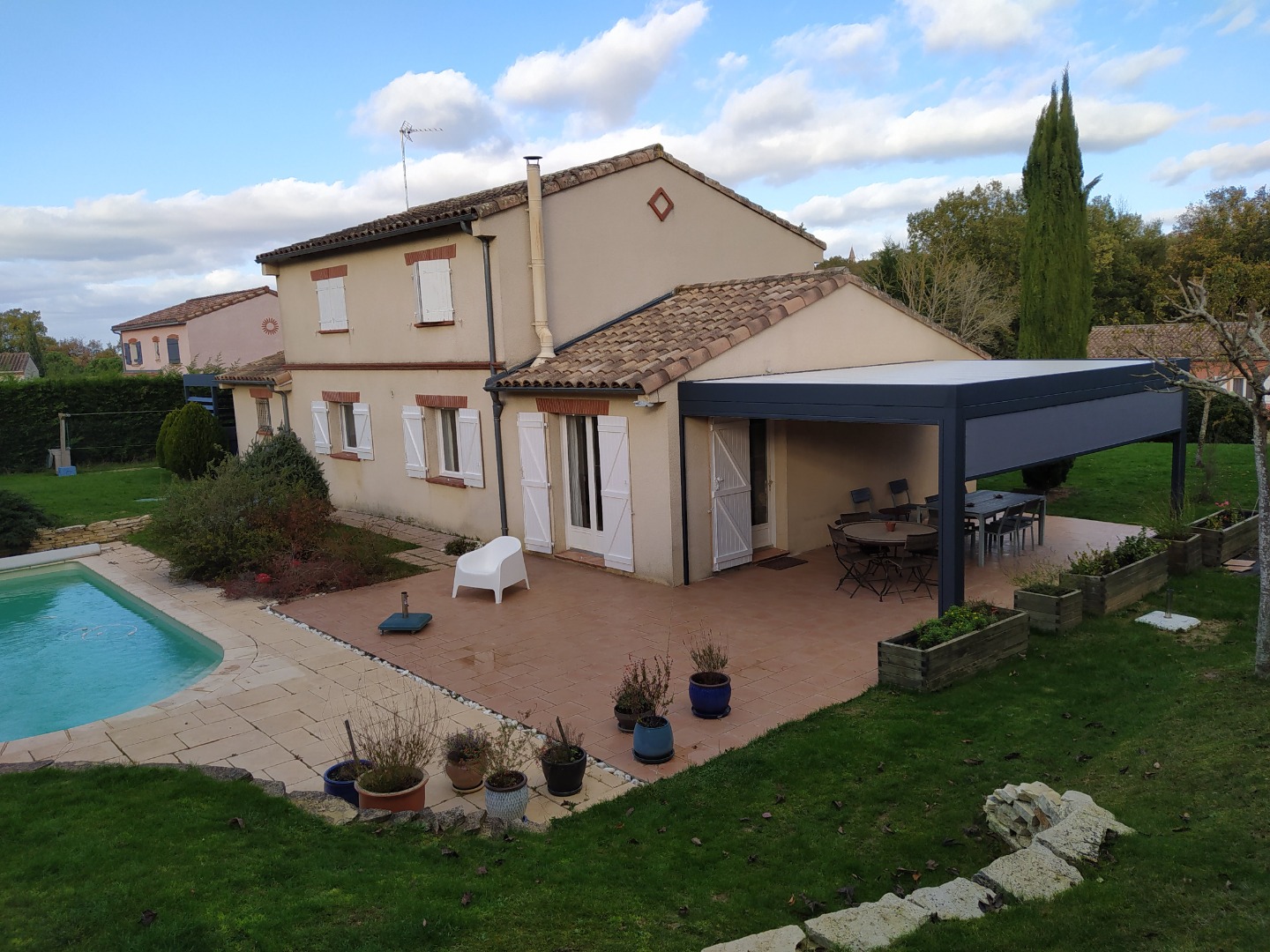 Maison méditerranéenne avec piscine, grande terrasse carrelée sous pergola grise, mobilier de jardin, et végétation luxuriante.